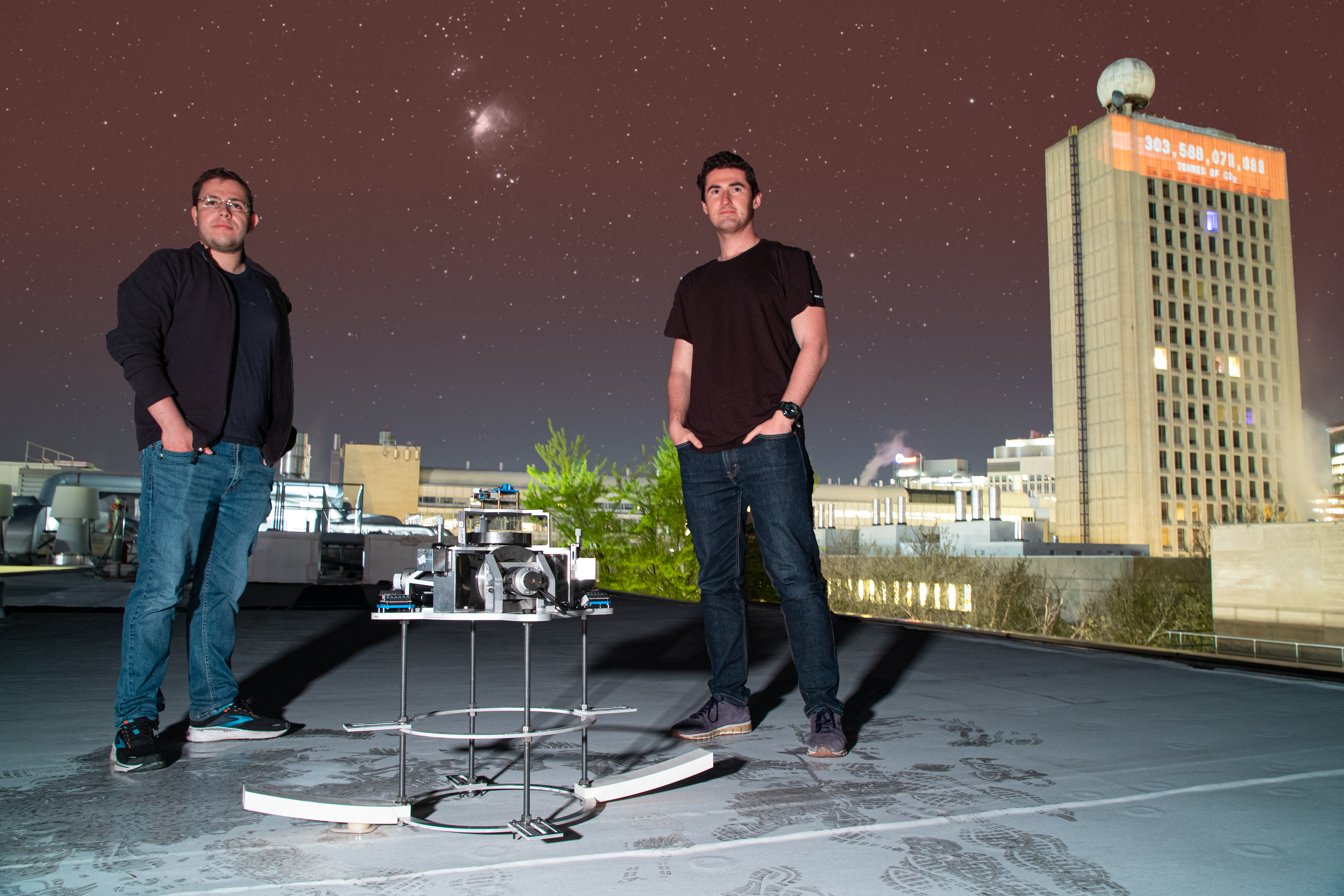 Alejandro Cabrales and Evan Kramer stand on a rooftop with the SpinAp space telescope hardware testbed between them. A purple/blue night sky can be seen in the background. To the right, a tall building is seen with an orange countdown clock illuminated toward the top with a number in the 300 billions appearing.