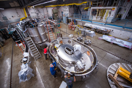 Three engineers working on the silver, donut-shaped magnet, placed inside a test stand