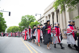 Procession of recent alumni wearing caps and gowns walks down the street.