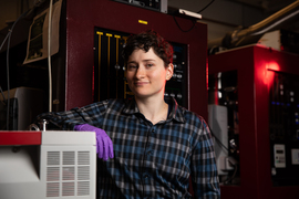 Alex Greene wears purple latex gloves with their arm on a grey machine, inside a crowded lab with large machines in background with knobs and sliders.