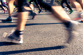 Low angle photo shows the legs and sneakers of about 12 runners. A blurry runner’s legs are in the foreground. 