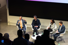 Joe HIggins, in a blue suit, speaks with a microphone to the audience while the other panelists, to his right, listen.