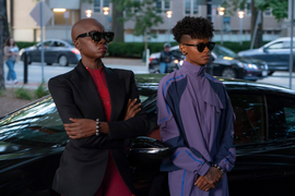 The two actors lean against a fancy car in an outdoor scene. 