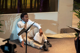 An actor wearing a tie sits on stage with a script in hand.