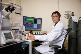 Guo, wearing a lab coat, sits at a desk in front of a computer inside a lab with large gas canisters and microscopes around him. The computer screen shows green and rainbow bars, and neon green circles.