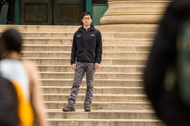 Guo stands on the steps of 77 Mass. Ave, and in the foreground are the blurred people framing the image.