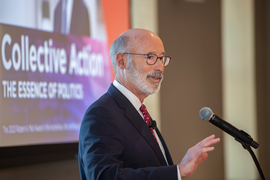 Tom Wolf speaks at the podium. In the background a screen shows the blurry words, “Collective Action.” 