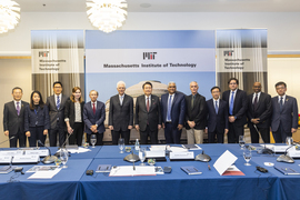 The 13 participants in the roundtable discussion stand side by side. In front of them is a wide table strewn with microphones, notepads, name cards, and water glasses. Behind them is a screen showing a photo of MIT’s Great Dome, with the MIT logo and the words “Massachusetts Institute of Technology.” 