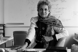 Monochrome photo of Cooper at desk with pens and markers. In the background is a whiteboard with text and boxy designs.