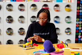 Sadler assembles Legos at a table, in the Lifelong Kindergarten group space. The background has a unique storage for Legos; they are inside fishbowl-style containers.