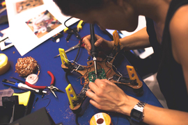 A person solders a circuit board on the underside of a drone-type device.