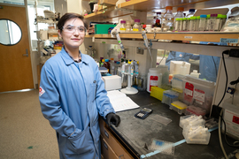 Farquhar wears a blue lab coat, black gloves, and safety goggles inside the lab. Their hand rests on a counter with lots of supplies, including vials and a calculator.