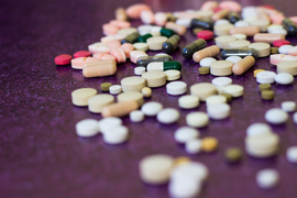 Cylindrical and round pills are scattered upon a purple surface with a vignette-like blur effect.