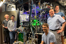 The 5 people stand around a large, crowded machine, full of dangling wires, green lights, blue LEDS. 