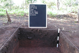 An archeological site is divided, by white rope, into a grid. One square has been dug out, and a sign says, “Xingu Nokugu… May 4, 2005. Midden”