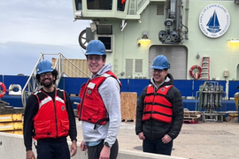 Three team members wear life jackets and hard hats while outside in a work area.