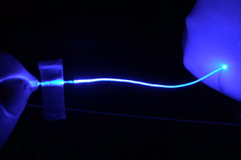 A blue glowing fiber in darkness. The fiber is held by finger and seems to light up with it touches another hand.