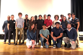 About 18 people, including teachers and students, stand on stage for a group photo.