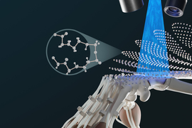 A robotic hand is being constructed by the printer. White spheres are in layers that are being cured by UV light beaming down from above. An inset shows the molecular makeup of the white spheres. 