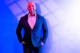 Justin Solomon stands in front of a wall and is lit with dramatic pink and blue light, with grid-like shadows on the wall.