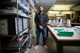 Baran Mensah stands in a studio filled with organized shelves and small rovers on large tables. Mensah wears a Brass Rat ring.