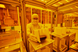 A person wearing head-to-toe PPE, including hood, gloves, and coveralls, stands at a small table inside an MIT.nano cleanroom. They are wrapping a sheet of plastic-like material around a circular form.