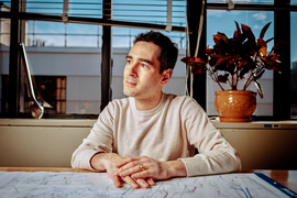 Rafael Jaramillo sits in his office and looks to the side. A large wrench sits on the window sill. The desk is covered in white paper with many drawings and notes on it.