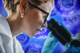 A lab researcher looking through a microscope with human cells in the background