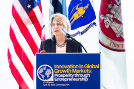 President Sally Kornbluth speaks at the podium, with flags in background.