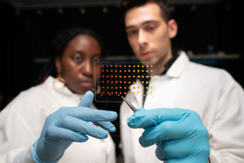 Eunice Aissi, left, and Alexander Siemenn each hold up a clear mat with dotted samples with gloved hand. They are wearing lab coats.