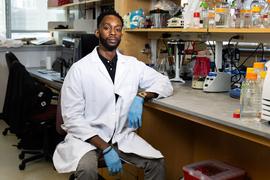 Feyisayo Eweje wears lab coat and gloves while sitting in a lab.