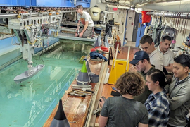 The busy tow tank lab has a big pool. Students mingle and observe a ship model on the water.