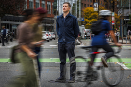 Andres Sevtsuk stands in the middle of a crosswalk as blurry travelers go by.