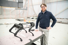 Luca Carlone smiles in a large room used for aerial drones, with many nets on the walls. He stands next to a table with a grey 4-legged robot with a “SPARK Lab” sticker.