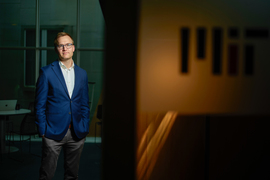 Kevin Dorst stands in a room in STATA. A blurry door in front bisects the photo and has the “MIT” logo.